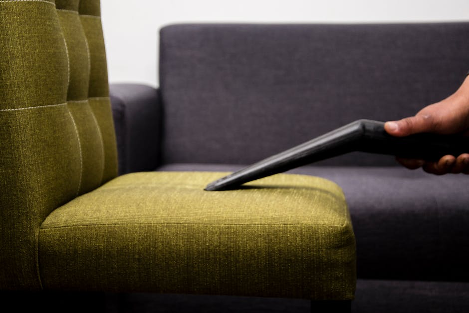 A person using a handheld electric upholstery cleaner on a dark gray fabric sofa in a residential living room, with visible cleaning process focused on the seat cushions. The sofa is placed near a whi